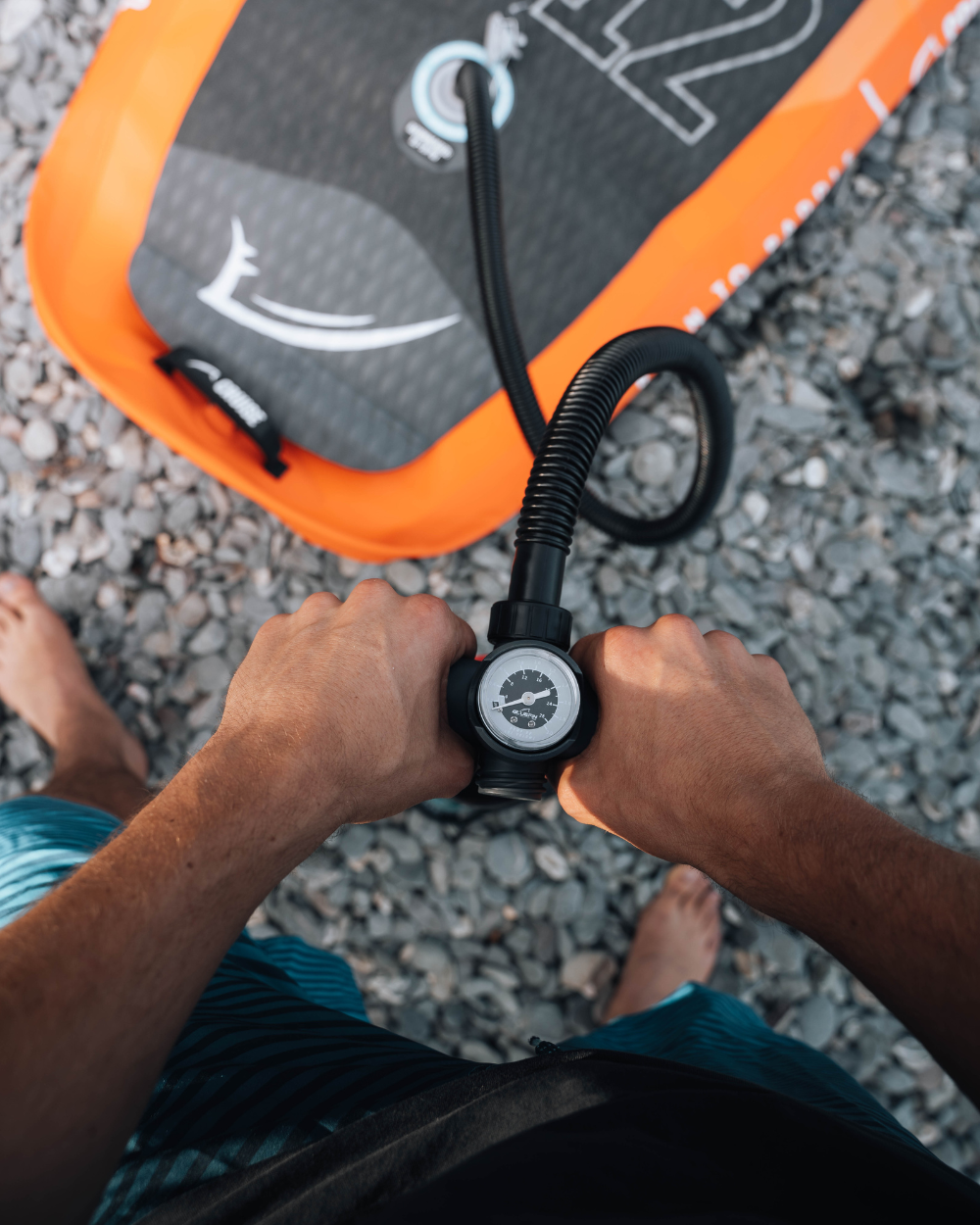 Person holding a black pump with pressure gauge, preparing to inflate an orange paddleboard on gravel.