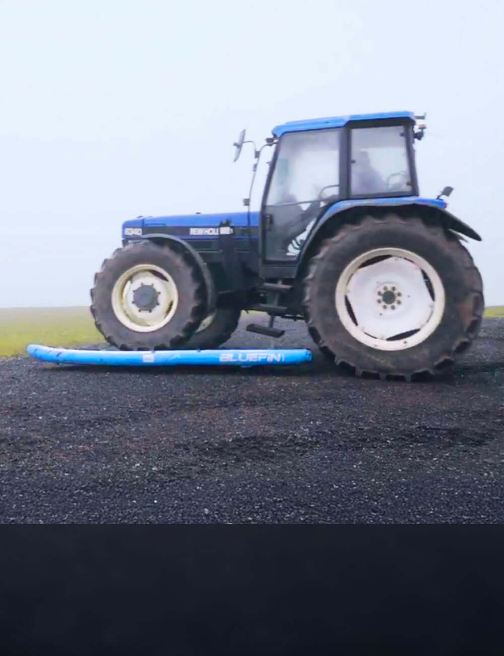 Blue inflatable paddleboard under a tractor tire on gravel ground in foggy weather