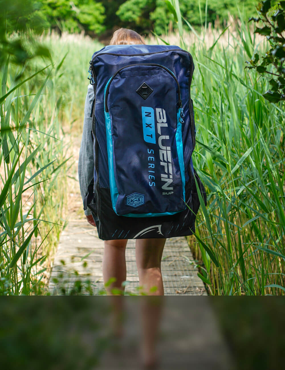 Person walking on a path carrying a large blue Bluefin NXT Series backpack surrounded by tall grass.