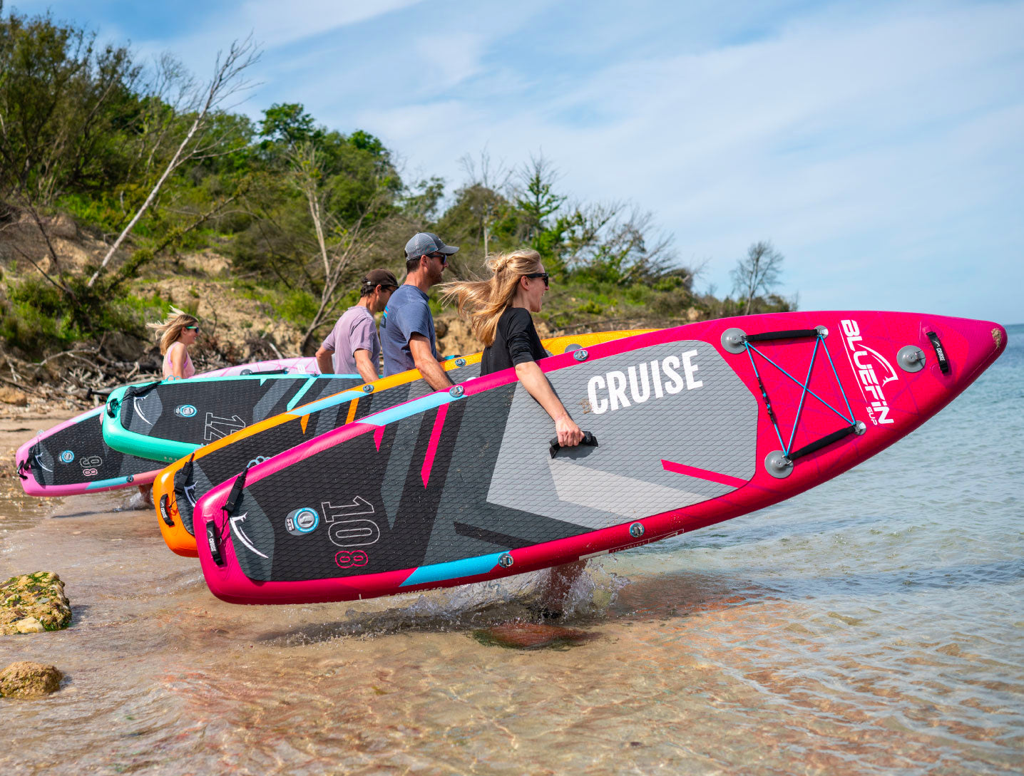 Four people carrying colorful inflatable paddleboards along a sandy shore by the water.