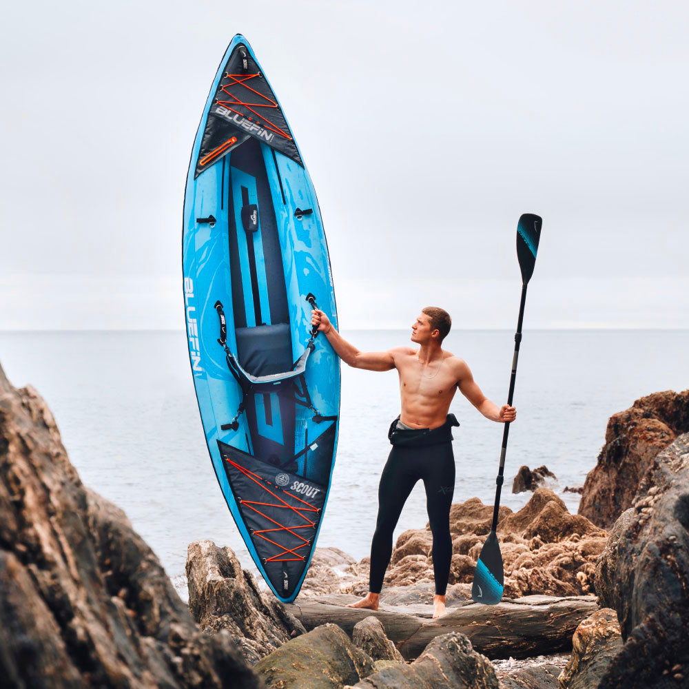 Man holding a blue inflatable paddleboard on rocky shore, wearing black wetsuit pants and no shirt.