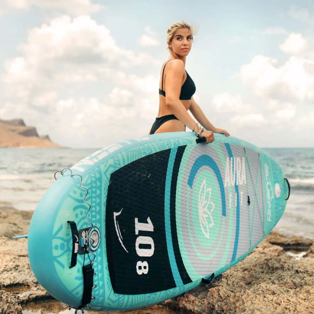 Frau in schwarzem Bikini, die ein türkisfarbenes aufblasbares Paddleboard am Ufer mit Ozeanwellen hält.
