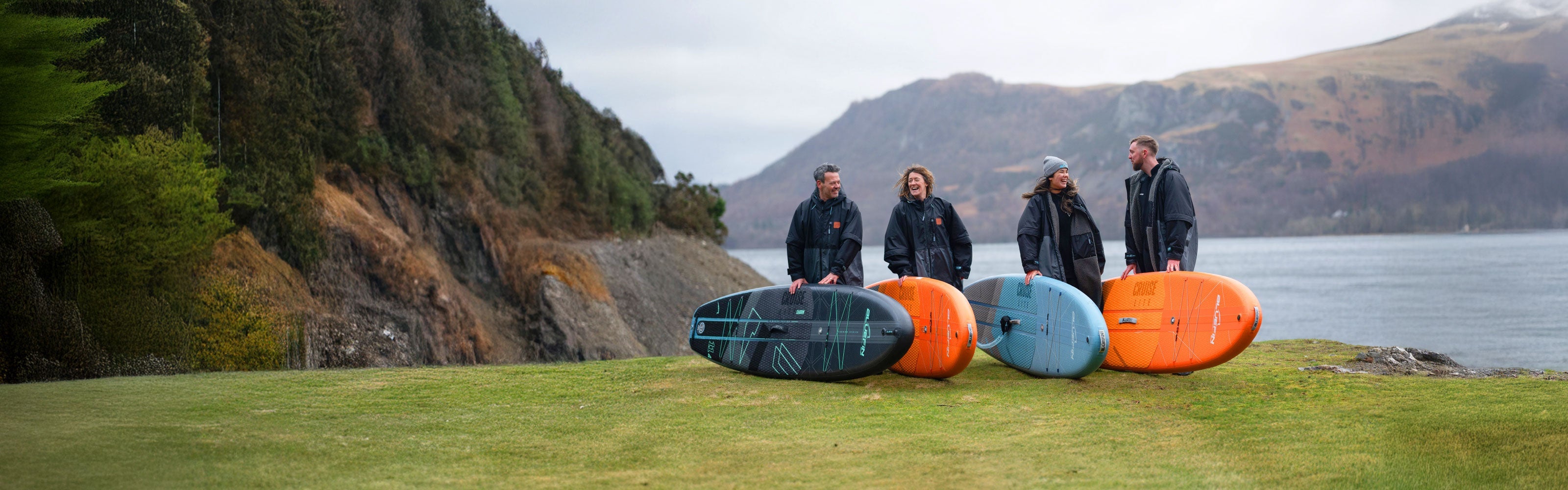 Four people standing on grass by a lake with colorful paddleboards in orange, blue, and black.