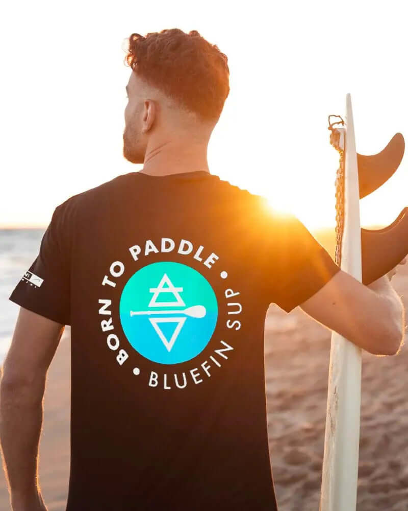 Persoon in een zwart T-shirt met 'Born to Paddle' en Bluefin SUP-logo, met een paddleboard in de hand.