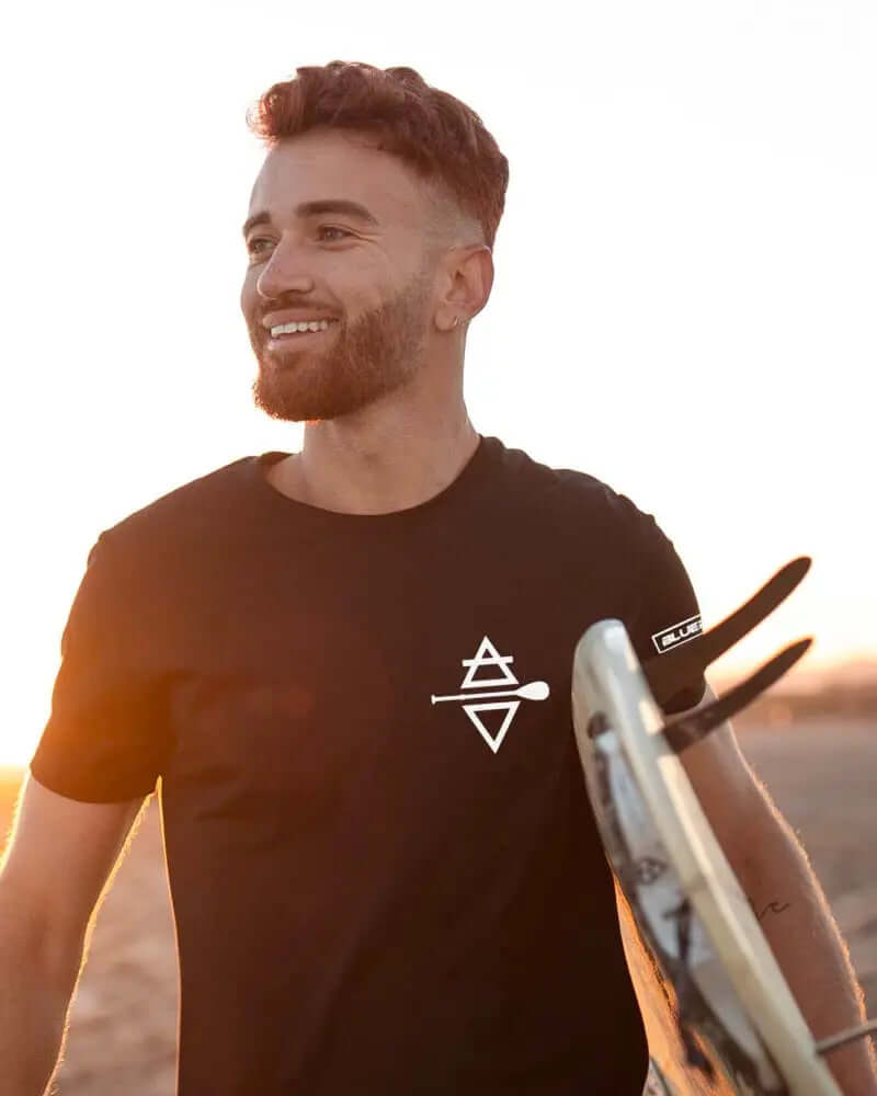 Man die glimlacht in een zwart T-shirt met logo, met een surfboard bij zonsondergang op het strand.