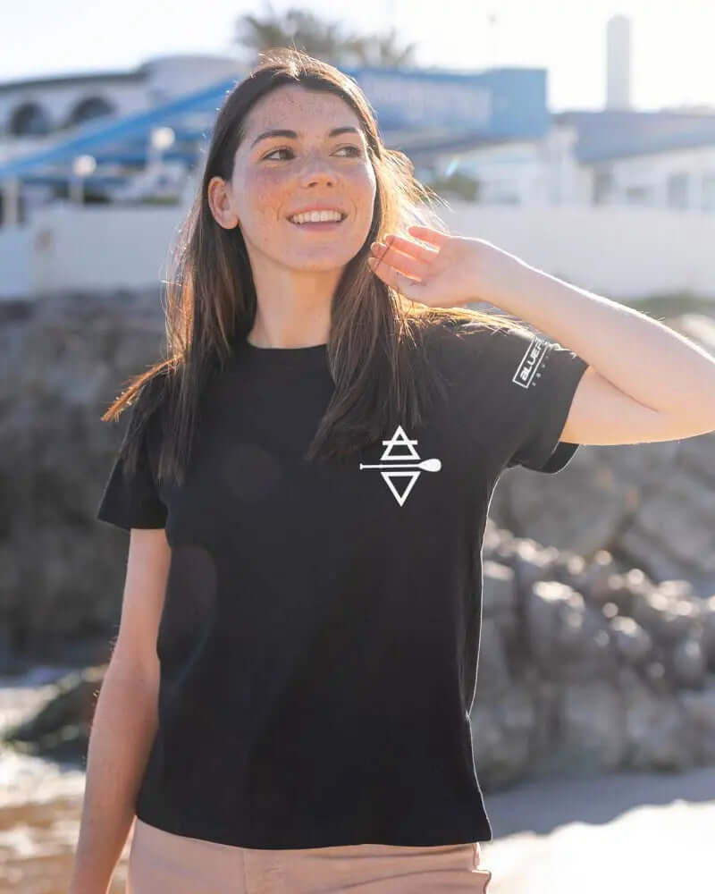 Vrouw in een zwart T-shirt met een wit logo, glimlachend en haar aanrakend bij het strand.