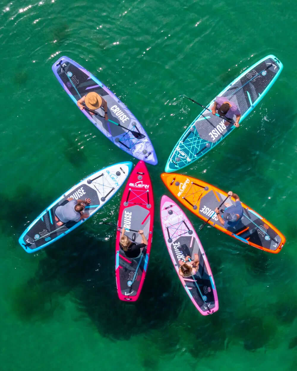 Six colorful paddleboards arranged in a circle on clear water, with people paddling on them.