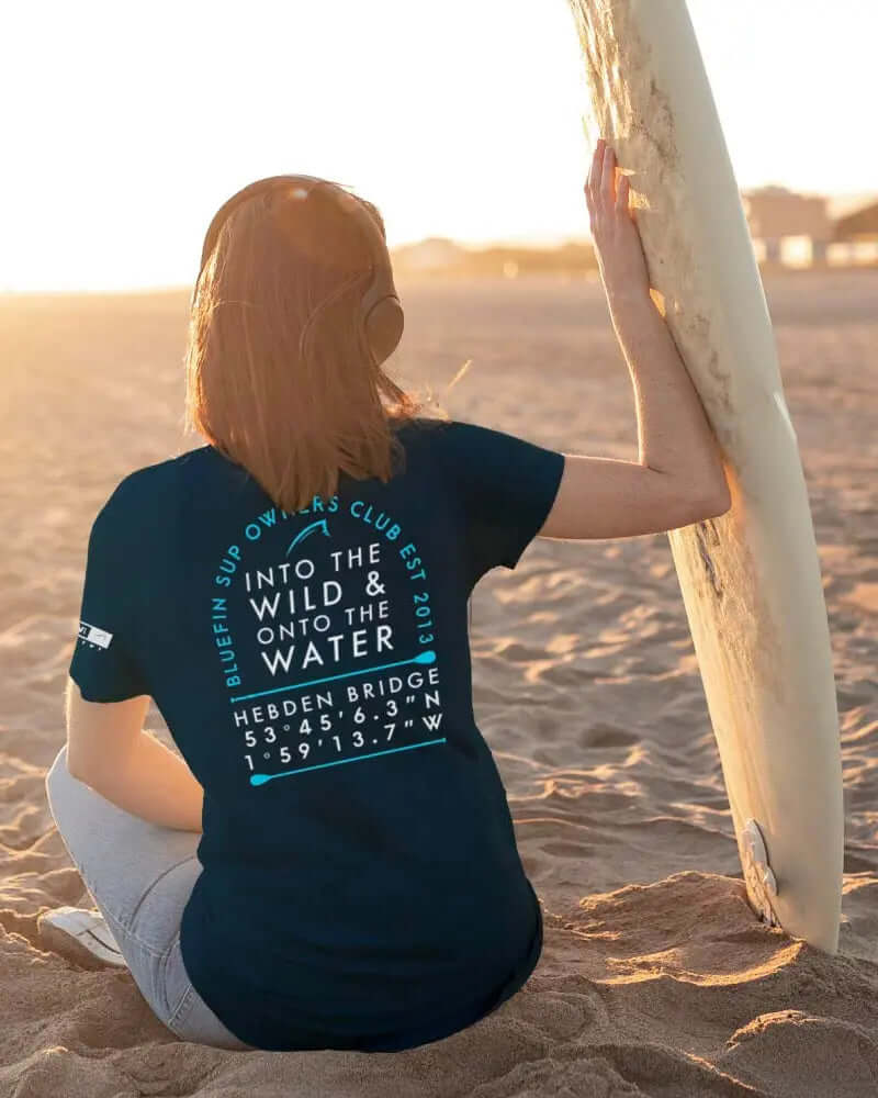 Persoon zittend op het zand in een marineblauw T-shirt met tekst en afbeeldingen, met een surfboard in de hand.