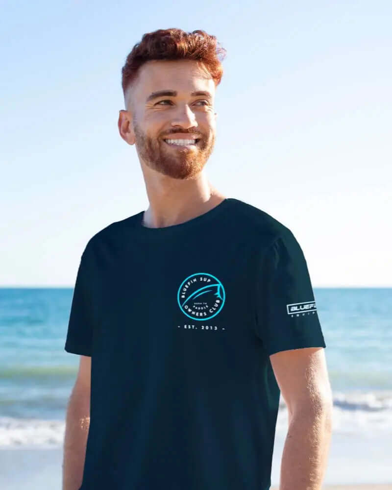 Man die glimlacht op het strand in een marineblauw Bluefin SUP T-shirt met logo op de voorkant en mouw.