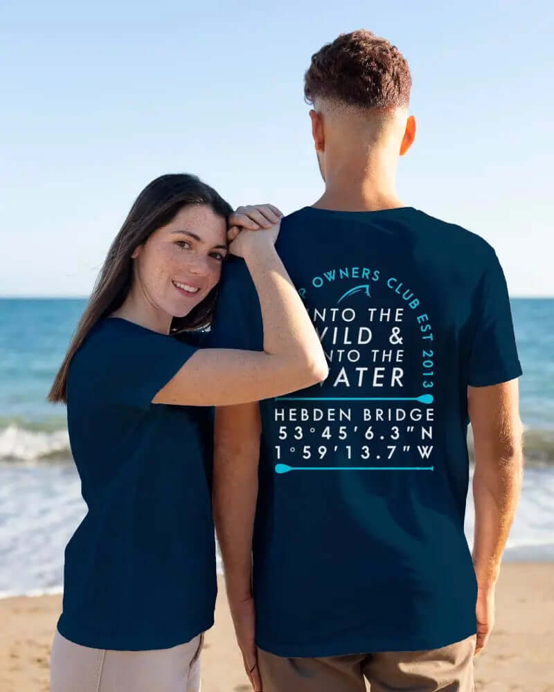 Koppel in bijpassende marineblauwe T-shirts met tekst op het strand, vrouw glimlachend en de schouder van de man aanrakend.