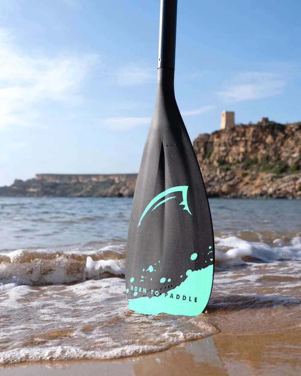 Black paddle blade with turquoise design, partially submerged in shallow water on a sandy beach.