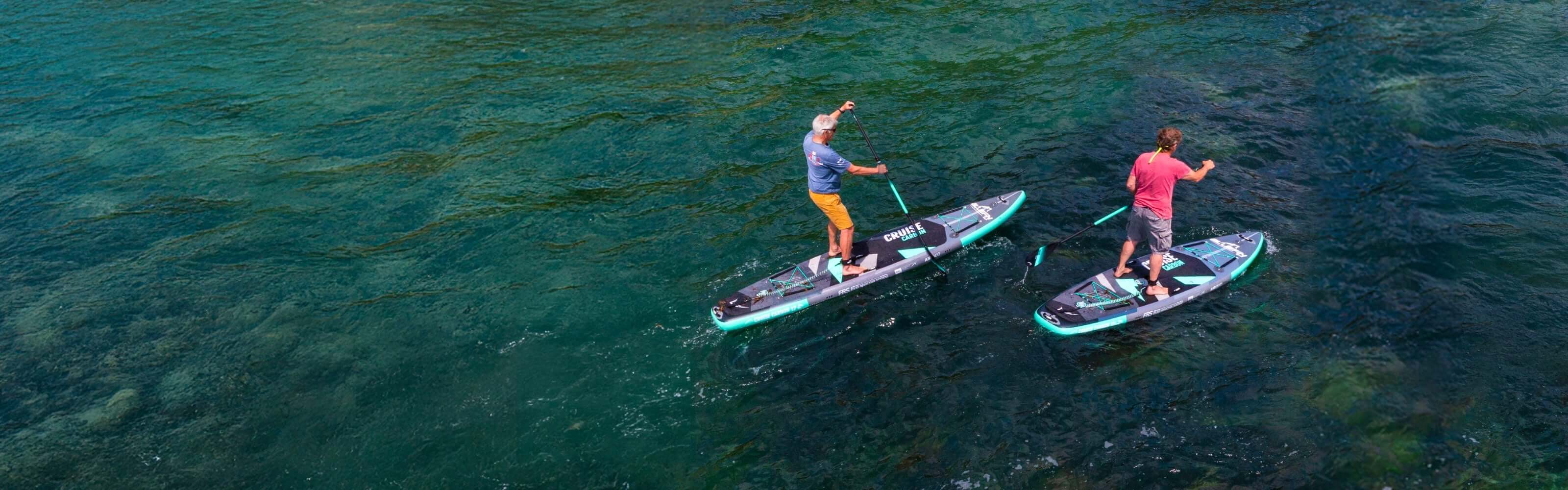 Two people paddleboarding on clear water, one in a blue shirt and orange shorts, the other in a red shirt.