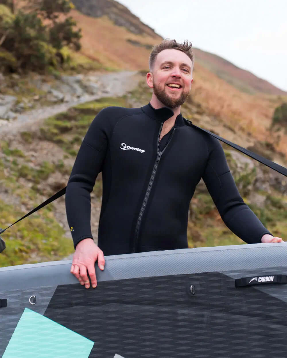 Man in a black wetsuit smiling while holding a paddleboard on a rocky path outdoors
