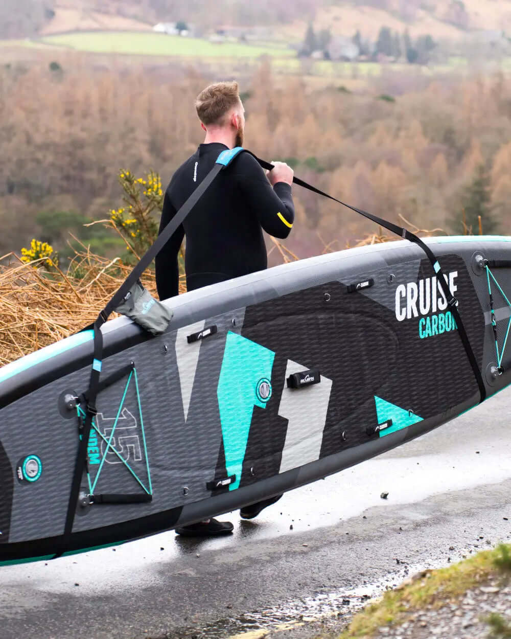Person in a wetsuit carrying a Cruise Carbon paddleboard along a road with trees in the background