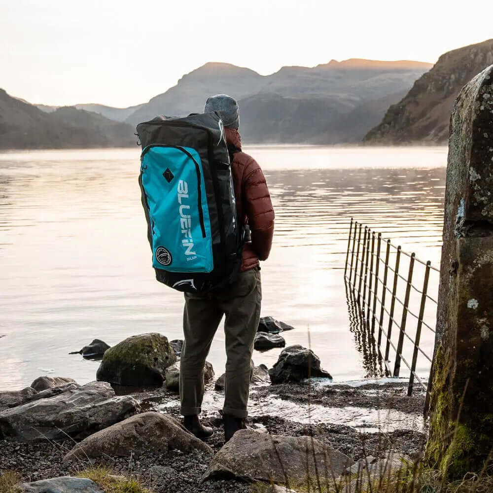 Persoon die bij een meer staat in een rode jas en grijze beanie, die een Bluefin SUP rugzak draagt.
