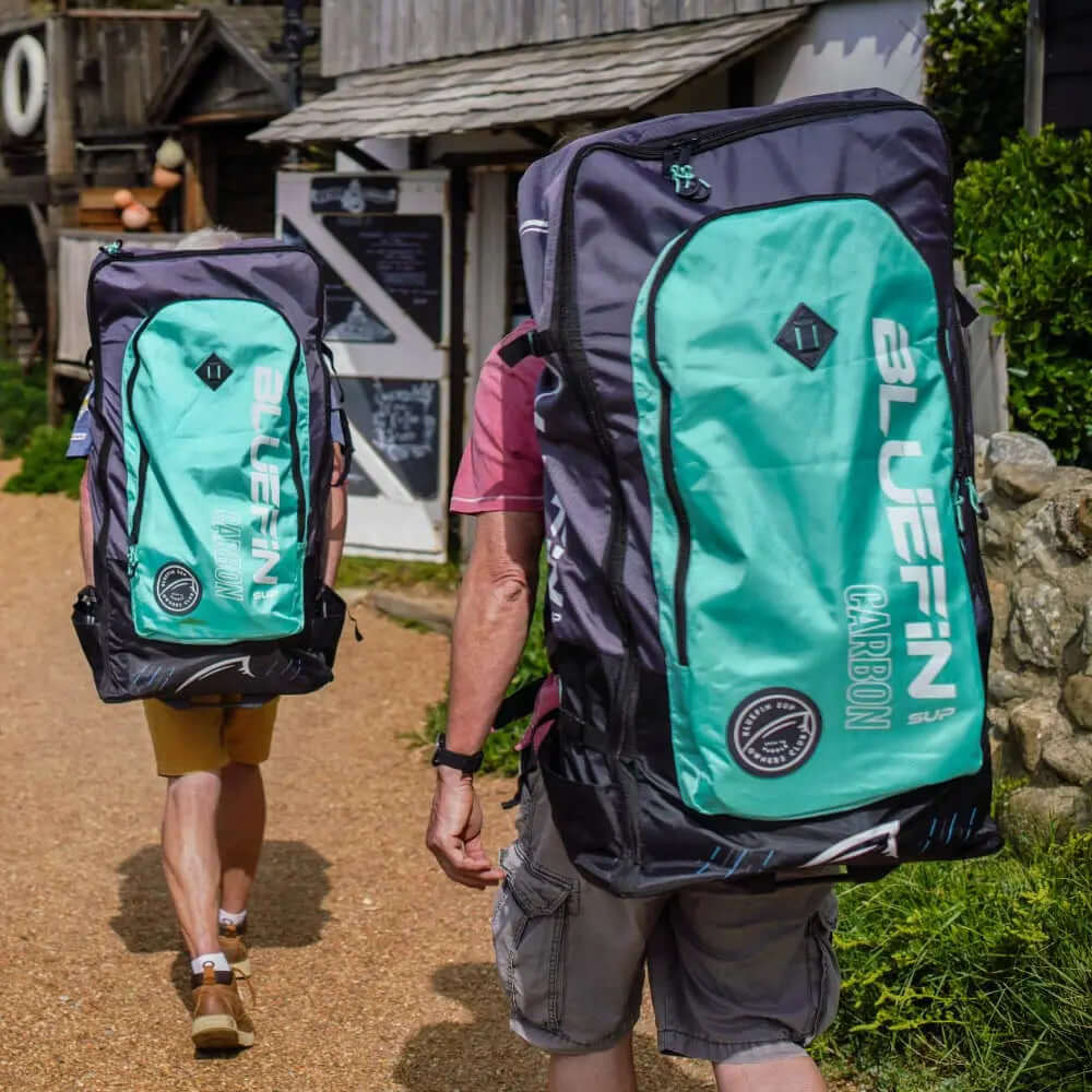 Deux personnes marchant avec de grands sacs à dos Bluefin Carbon SUP sur un chemin de gravier près de bâtiments.