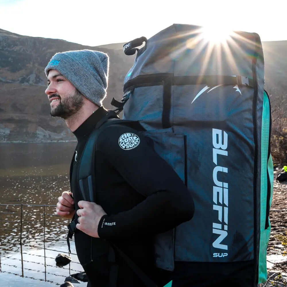 Homme en combinaison de plongée portant un grand sac à dos Bluefin SUP près d'un lac avec des montagnes en arrière-plan.