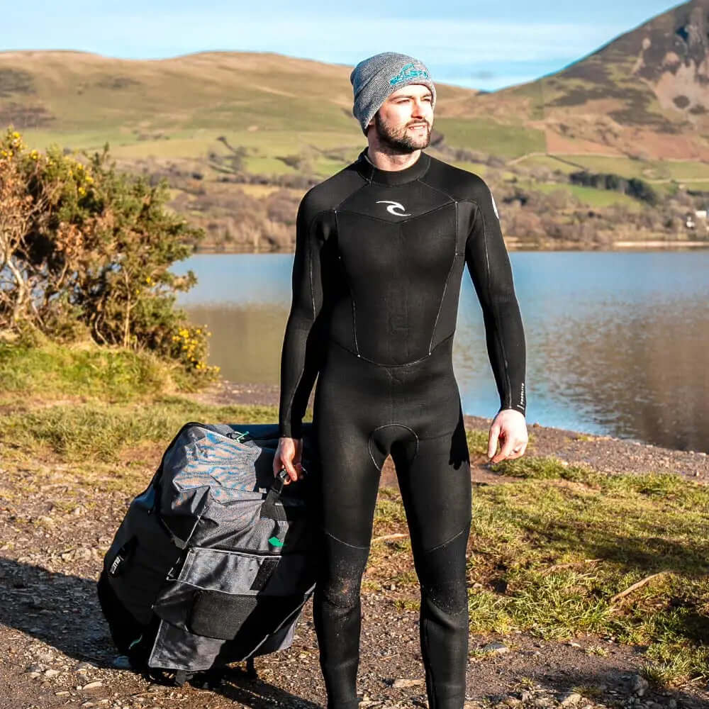 Homme en combinaison de plongée noire portant un sac à roulettes près d'un lac avec des collines en arrière-plan.