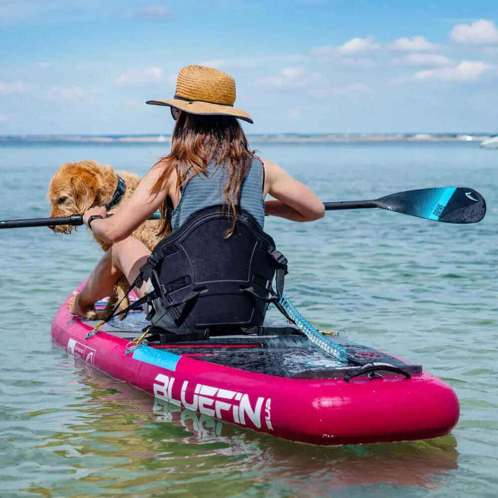 Person paddling a pink inflatable paddleboard with a dog, wearing a straw hat, in clear water