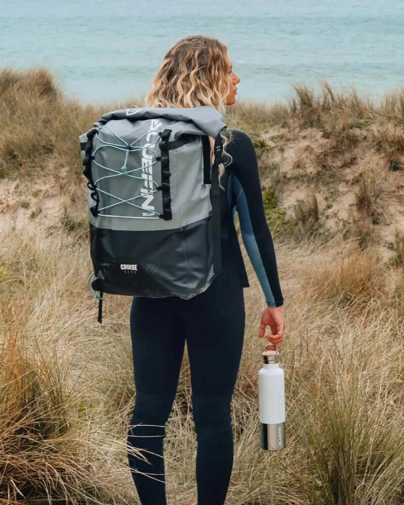 Persoon in een wetsuit die een waterfles vasthoudt, met een grijze rugzak van Bluefin SUP bij het strand.