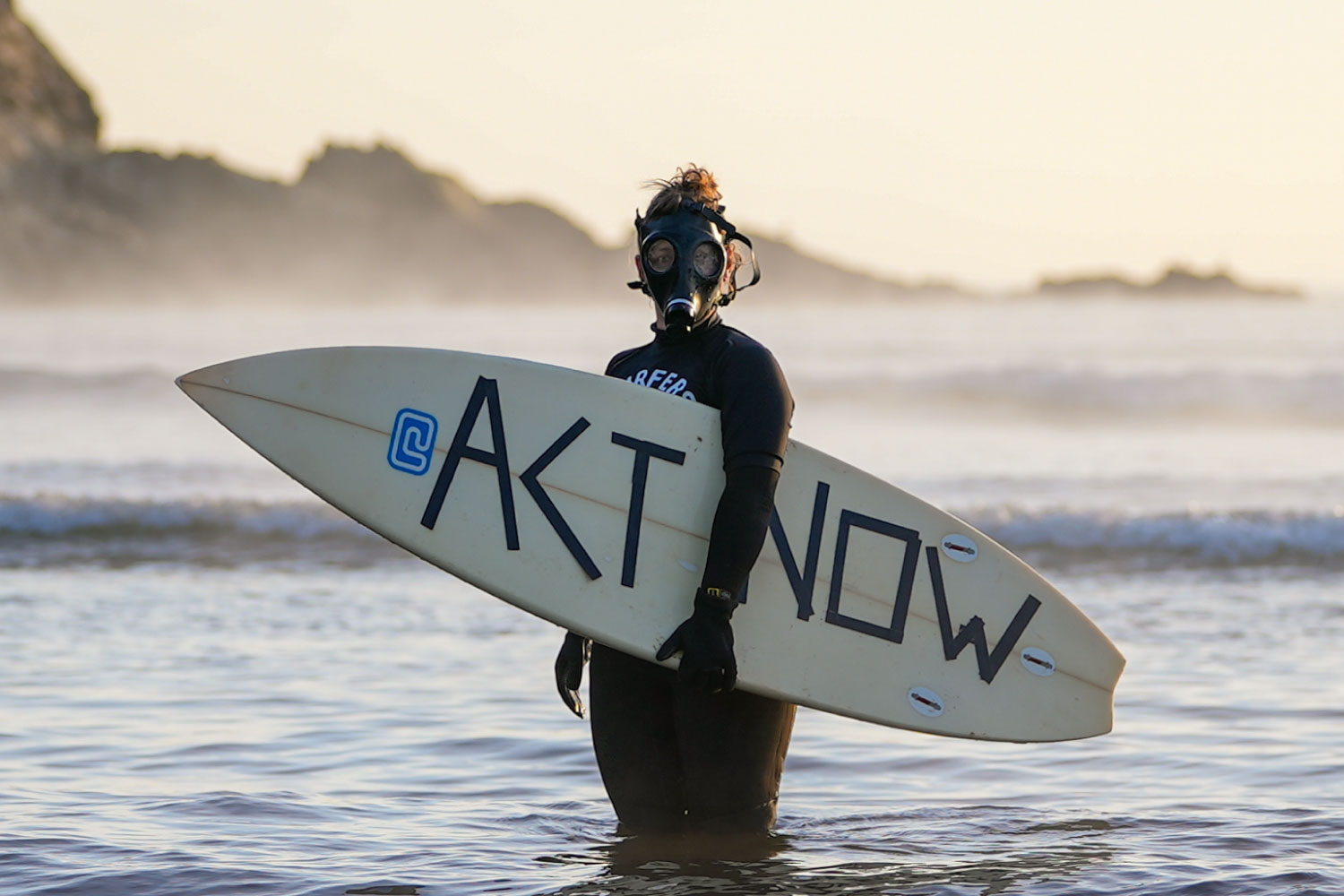 Surfer in a wetsuit and gas mask holding a surfboard with 'ACT NOW' written on it in shallow water.