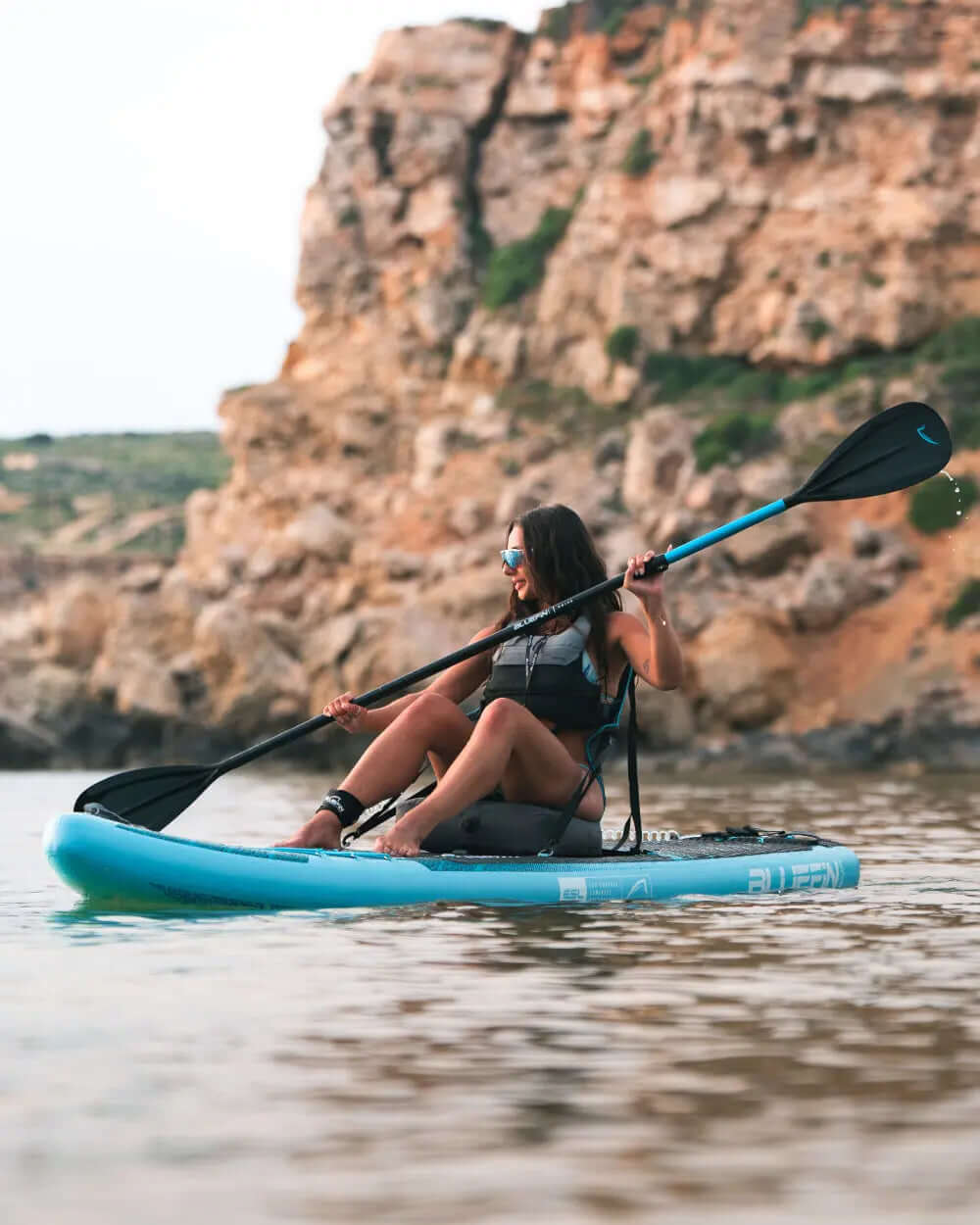 Vrouw die peddelt op een blauwe opblaasbare paddleboard nabij rotsachtige kliffen in kalm water