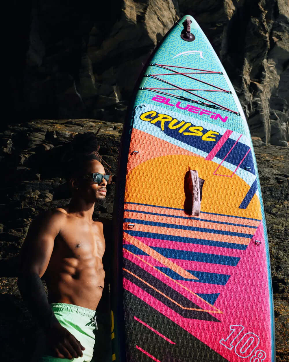 Colorful inflatable paddleboard with textured surface, center fin, and a man in sunglasses beside it