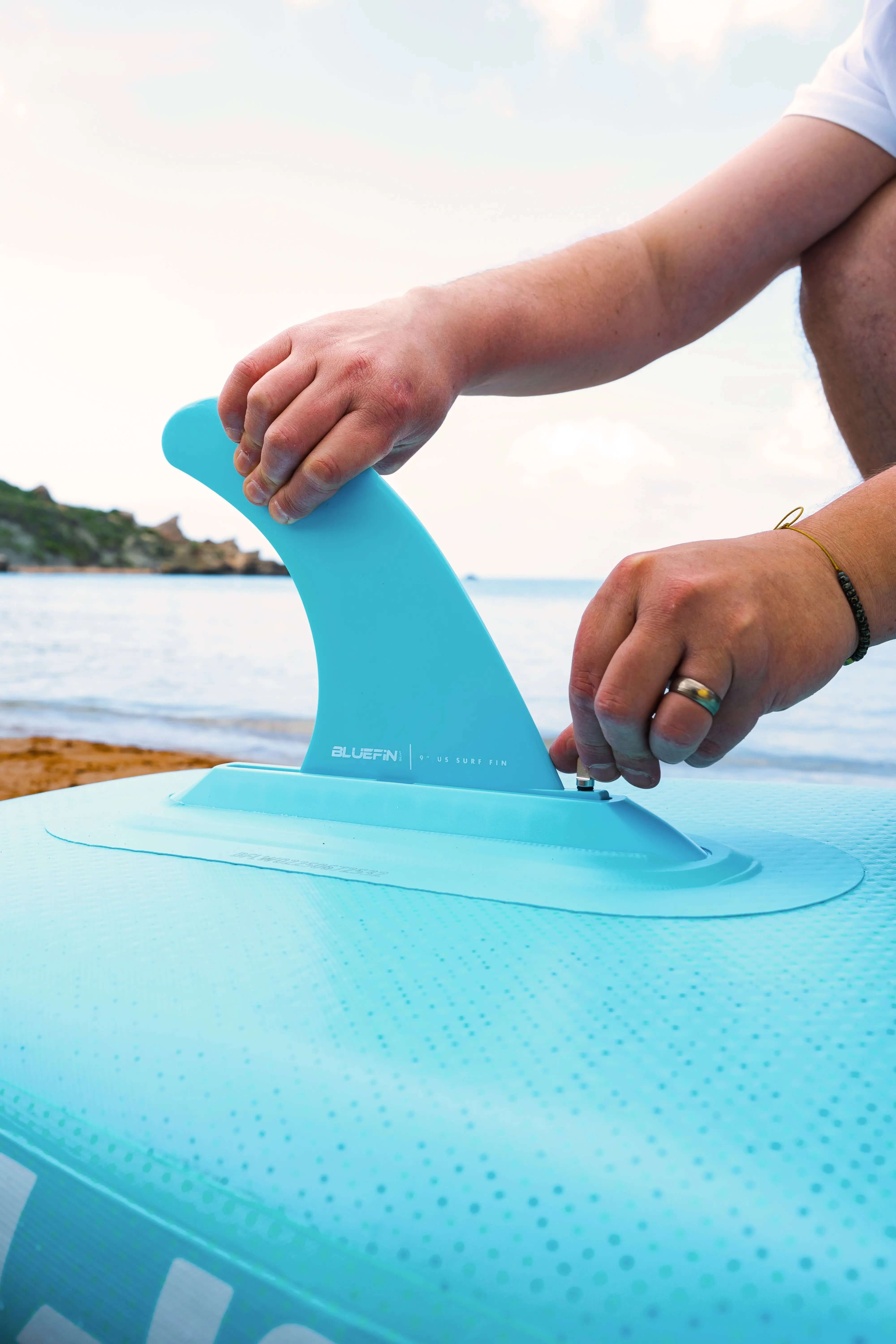 Person attaching a blue fin to a light blue inflatable paddleboard on a sandy beach