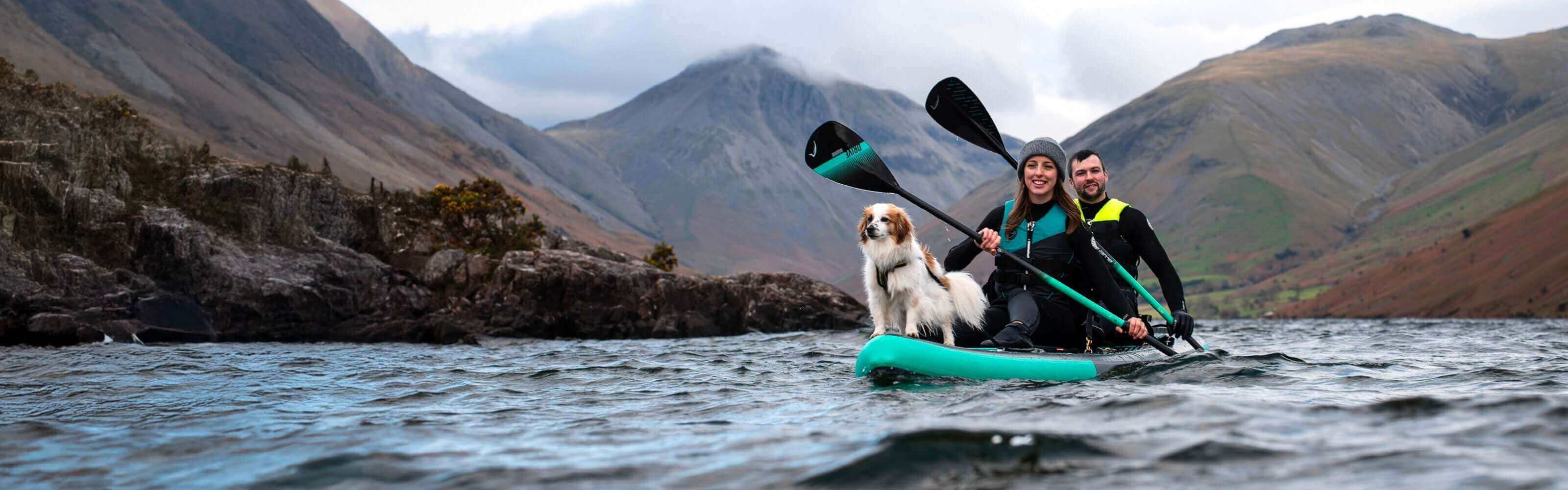 Two people and a dog on a teal inflatable paddleboard in a lake surrounded by mountains