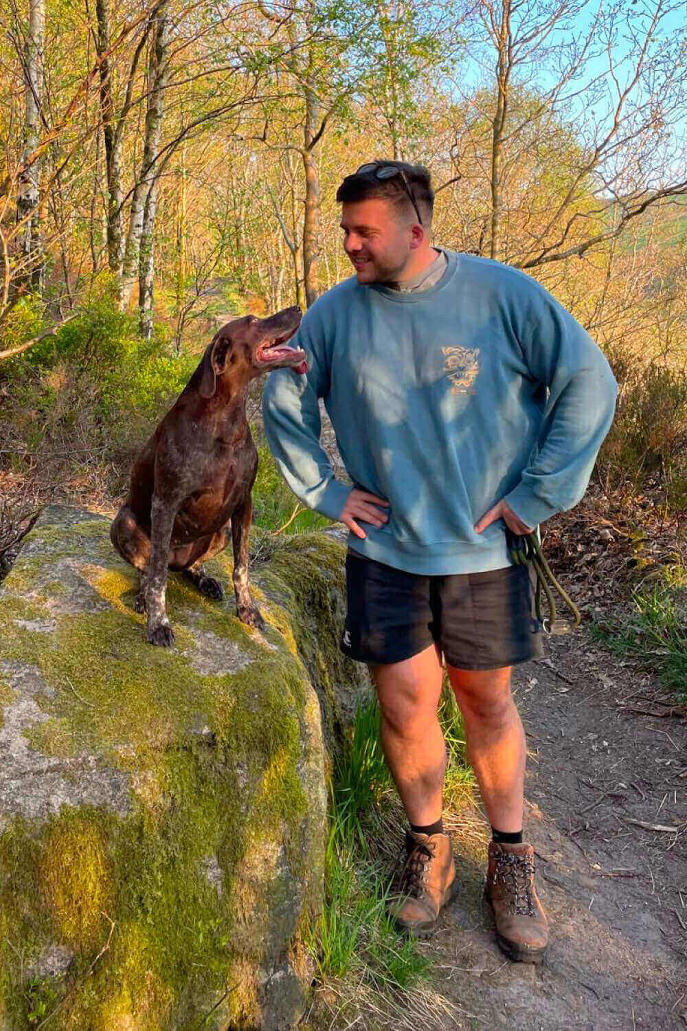 Man in a blue sweatshirt and shorts stands beside a brown dog on a mossy rock in a forest.