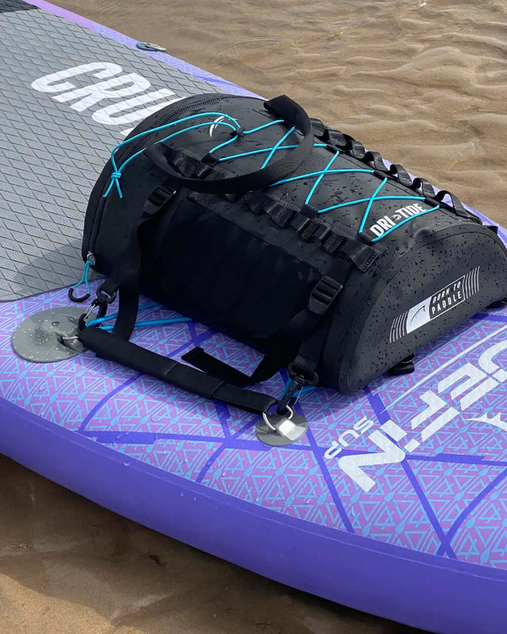 Black waterproof duffel bag with blue accents on a purple inflatable paddleboard on sandy beach