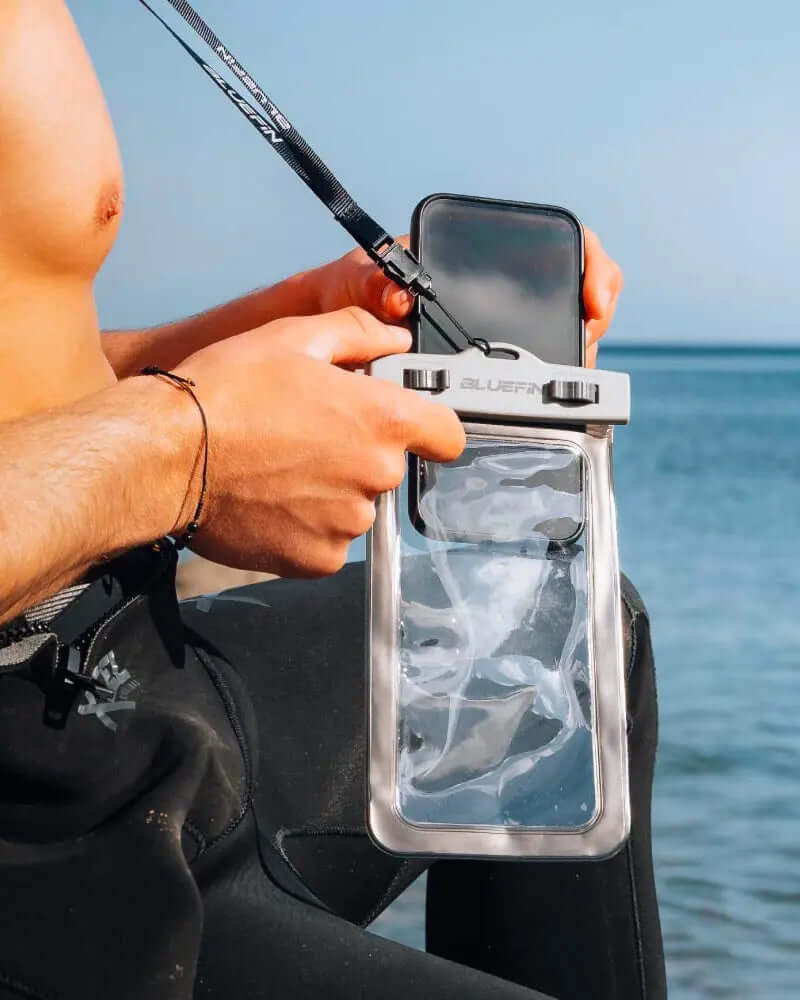 Persona sosteniendo una funda de teléfono impermeable con un cordón cerca del agua, vistiendo un traje de neopreno.