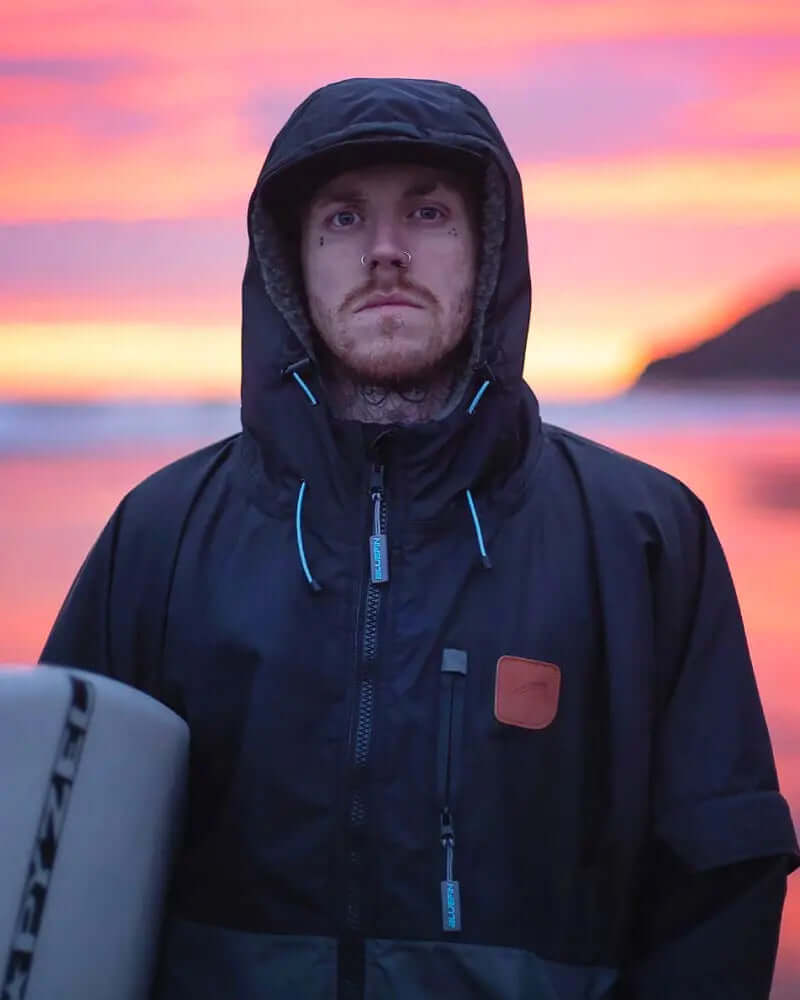 Persoon in een zwarte hoodie-jack die bij de oceaan staat tijdens zonsondergang, met een surfplank naast zich.