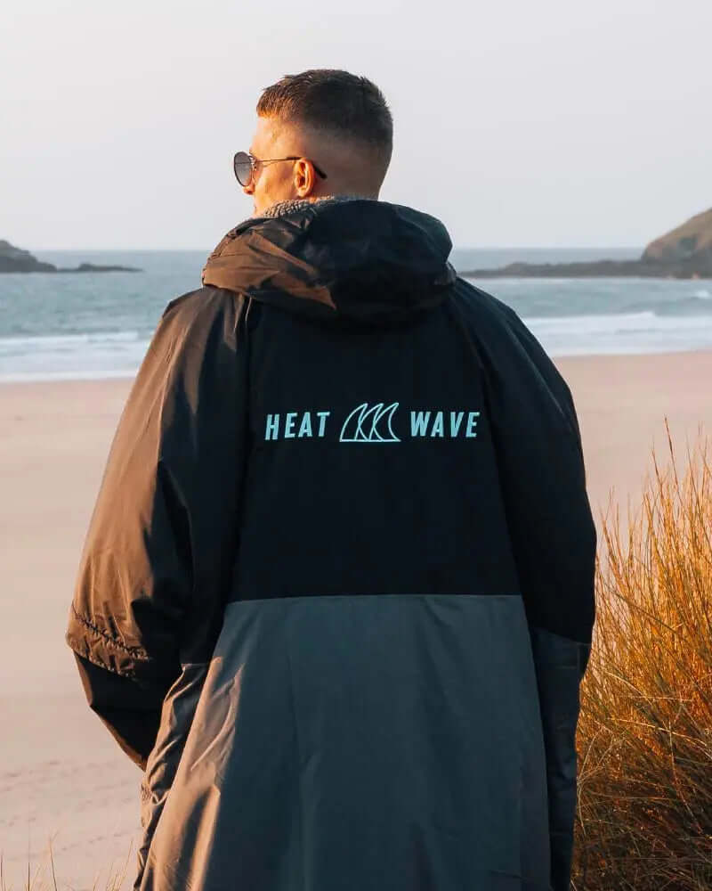 Persoon in een zwart-grijs jack met 'HEAT WAVE' logo, staande op een strand tijdens zonsondergang.