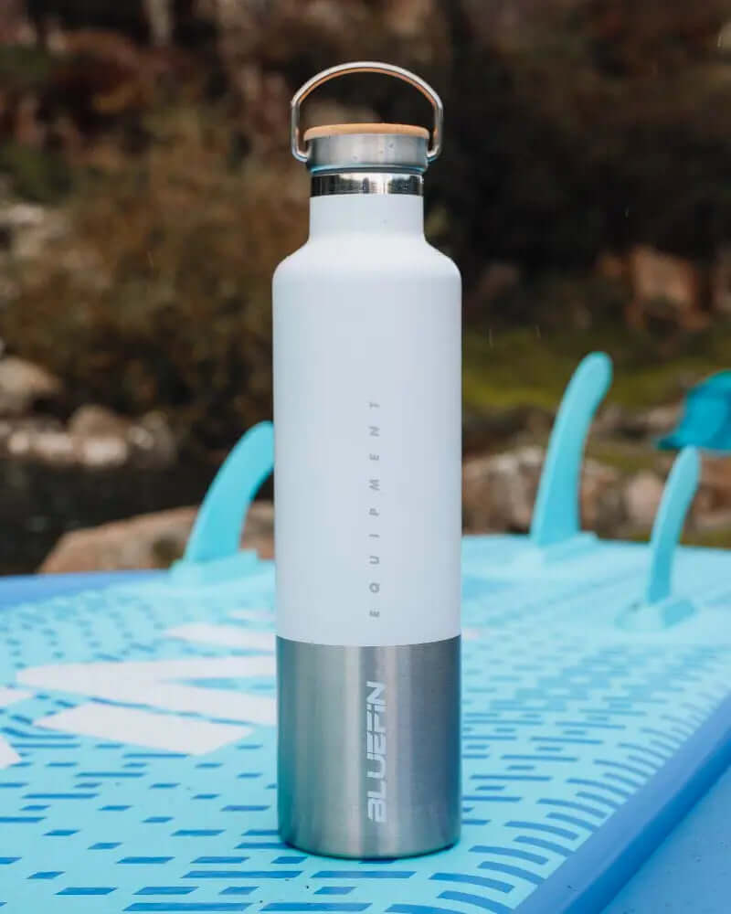 Botella de agua aislada blanca con fondo de acero inoxidable, colocada sobre una tabla de paddle azul.