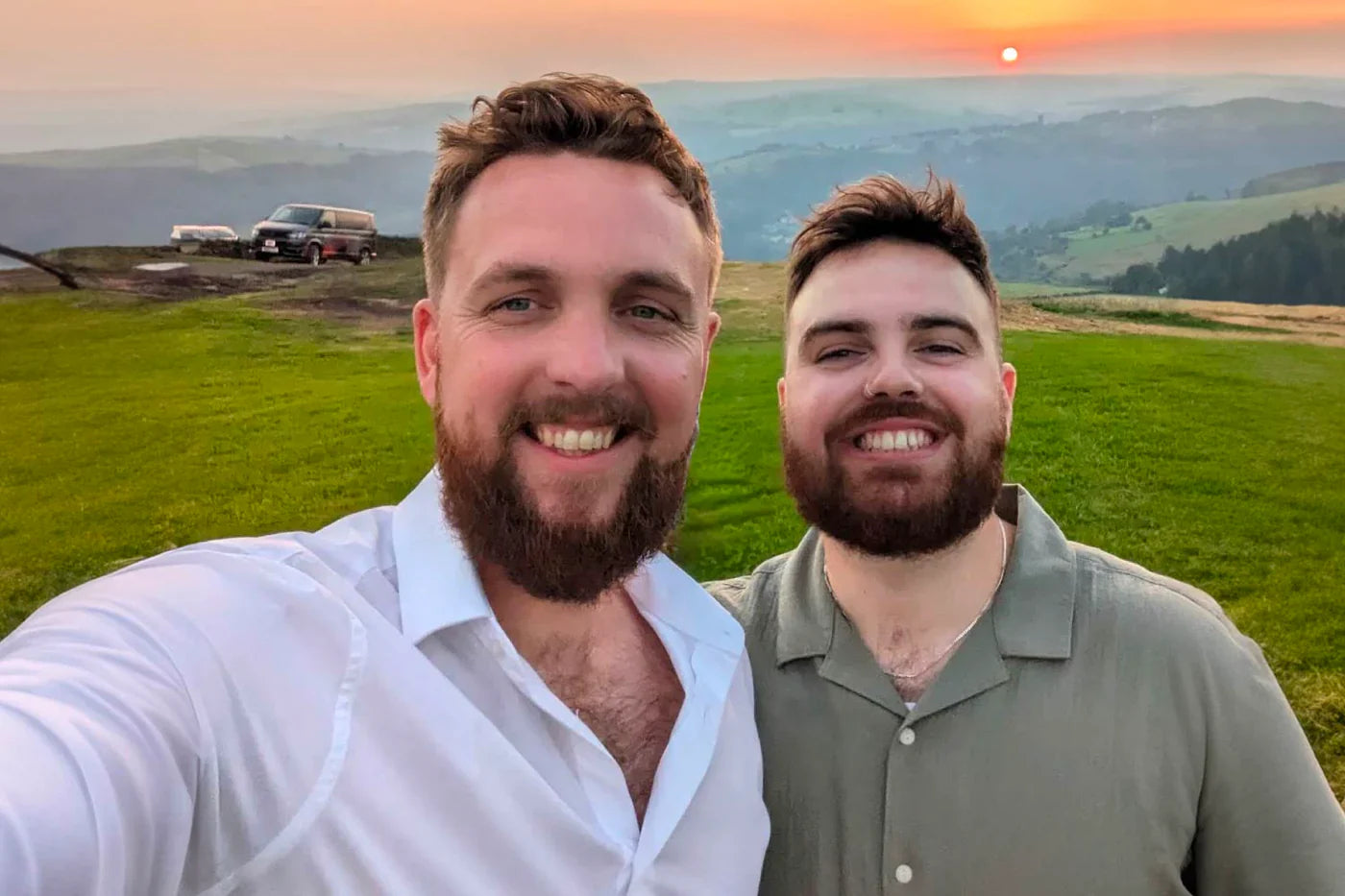 Two men smiling for a selfie on a grassy hilltop at sunset, with hills in the background.