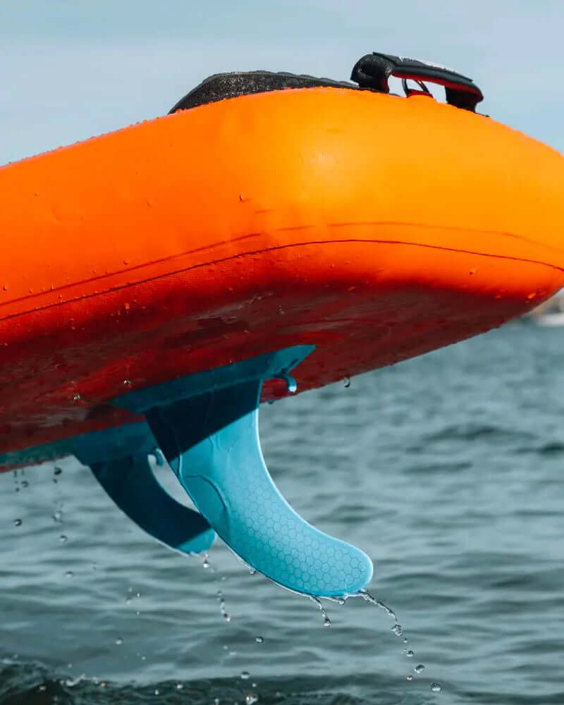 Paddle gonflable orange avec aileron bleu partiellement immergé dans l'eau, gouttes éclaboussant autour.