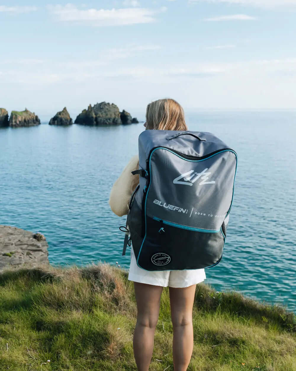 Person standing on grass by the water, wearing a white outfit and carrying a large Bluefin backpack.
