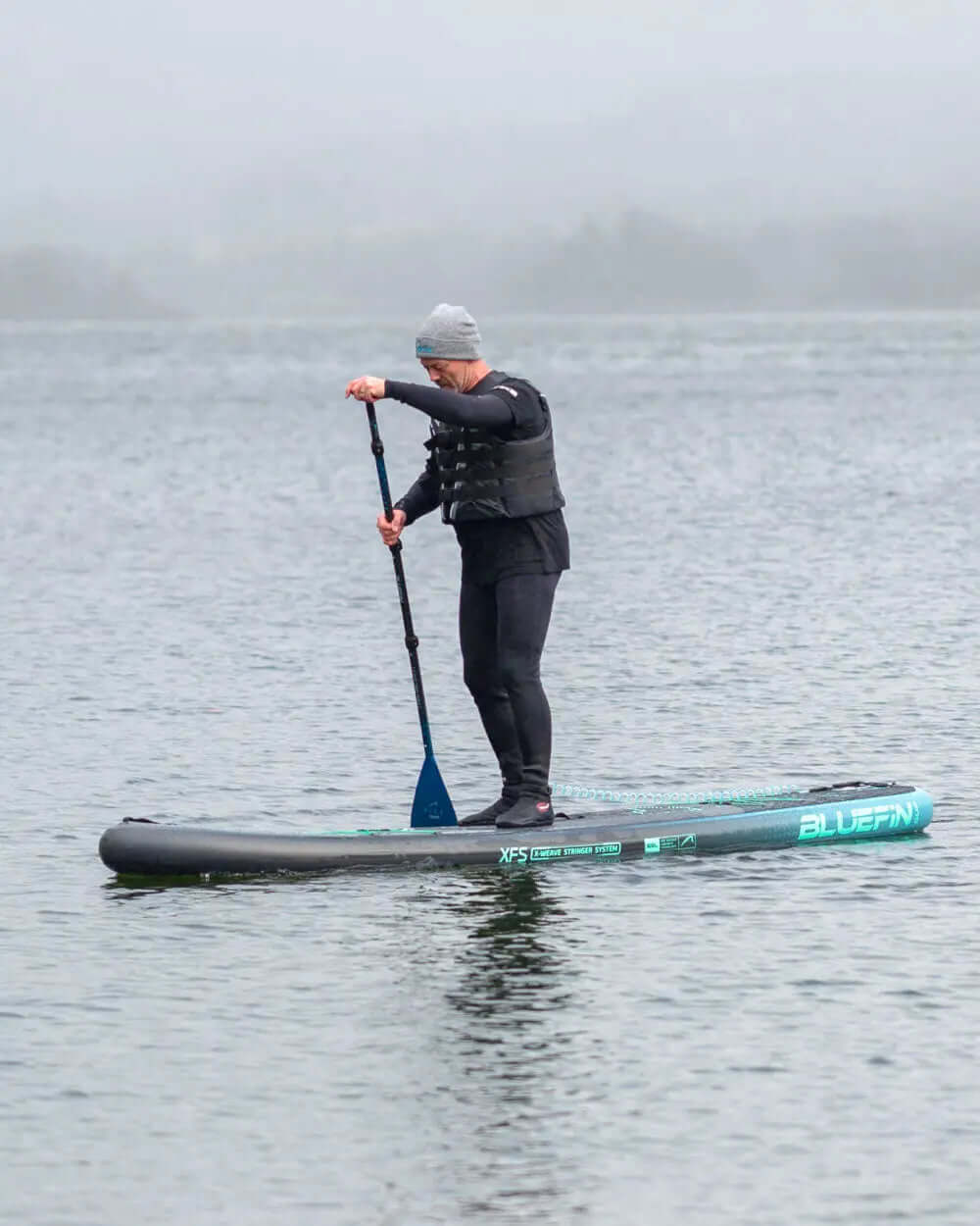 Person paddling on a Bluefin inflatable paddleboard in calm water, wearing a life vest and beanie.