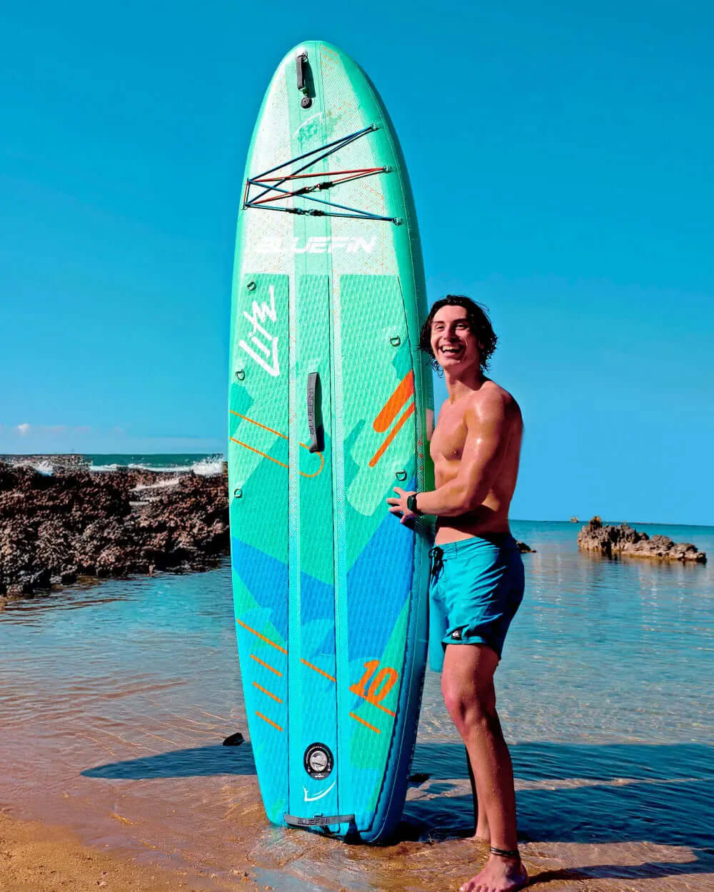 Man smiling while holding a turquoise paddleboard on a beach with clear blue sky and water.