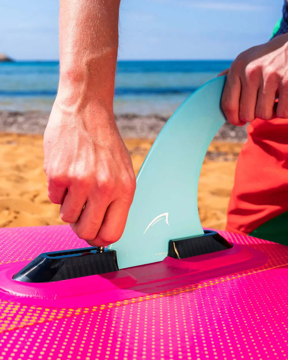Person attaching a blue fin to a pink inflatable paddleboard on a sandy beach