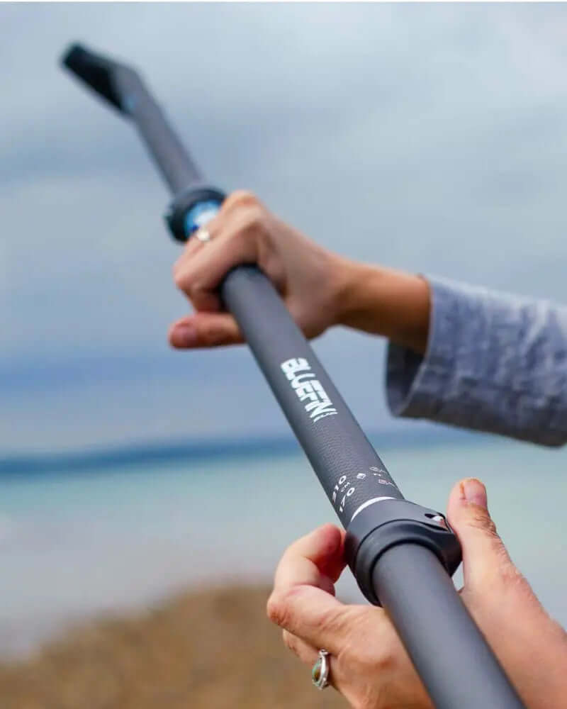 Persoon houdt een zwarte verstelbare peddel met een gestructureerde grip en een gebogen uiteinde, tegen een bewolkte lucht.