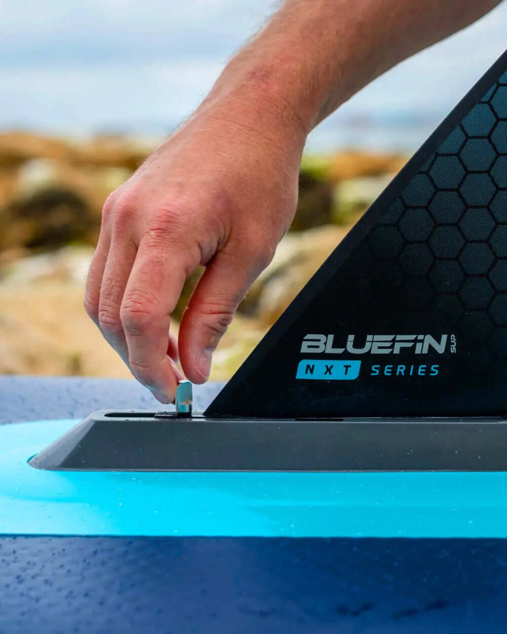 Hand securing the fin on a Bluefin SUP NXT Series inflatable paddleboard by the water