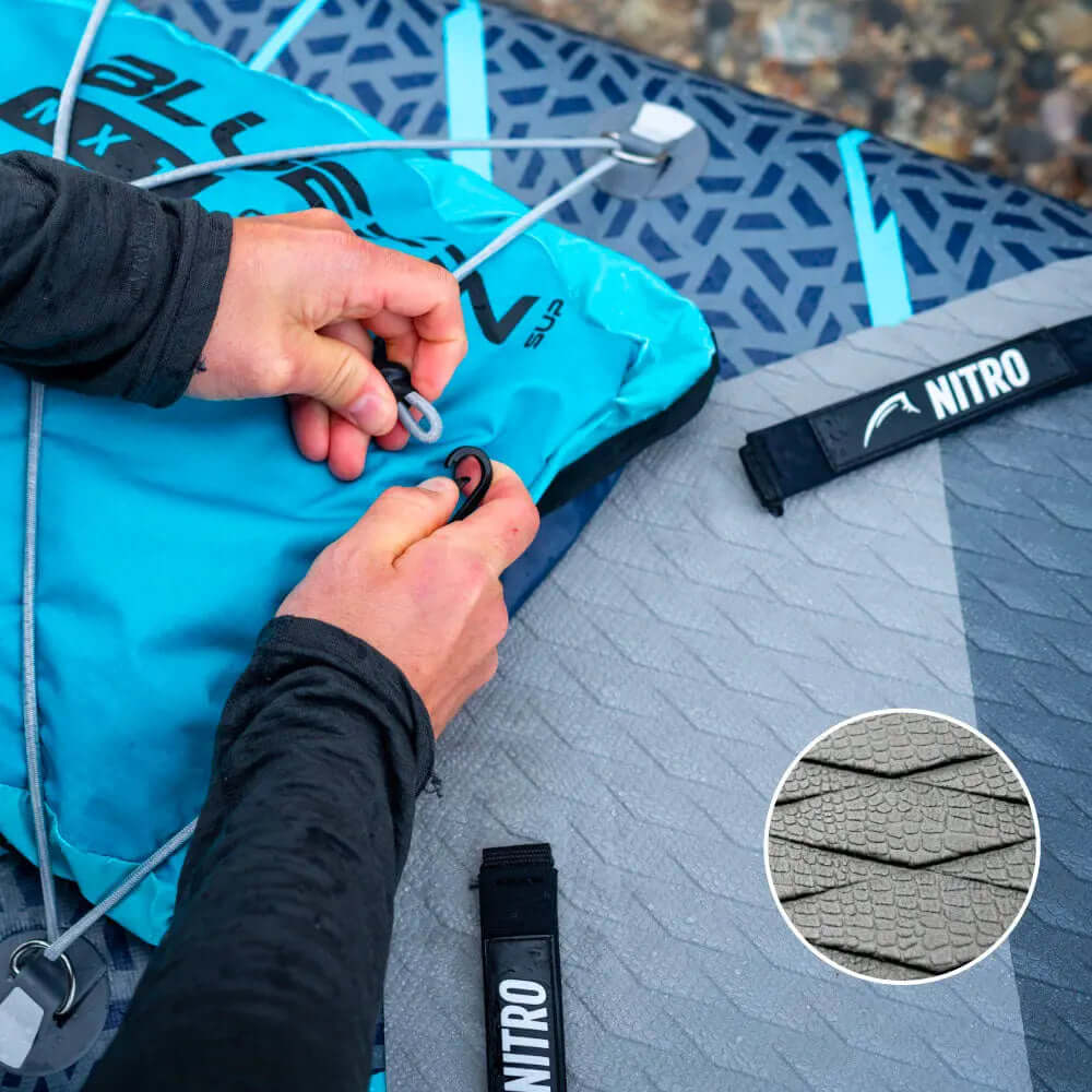 Hände, die eine blaue aufblasbare Paddleboard-Tasche mit einer grauen strukturierten Deckplatte und NITRO-Gurt sichern.