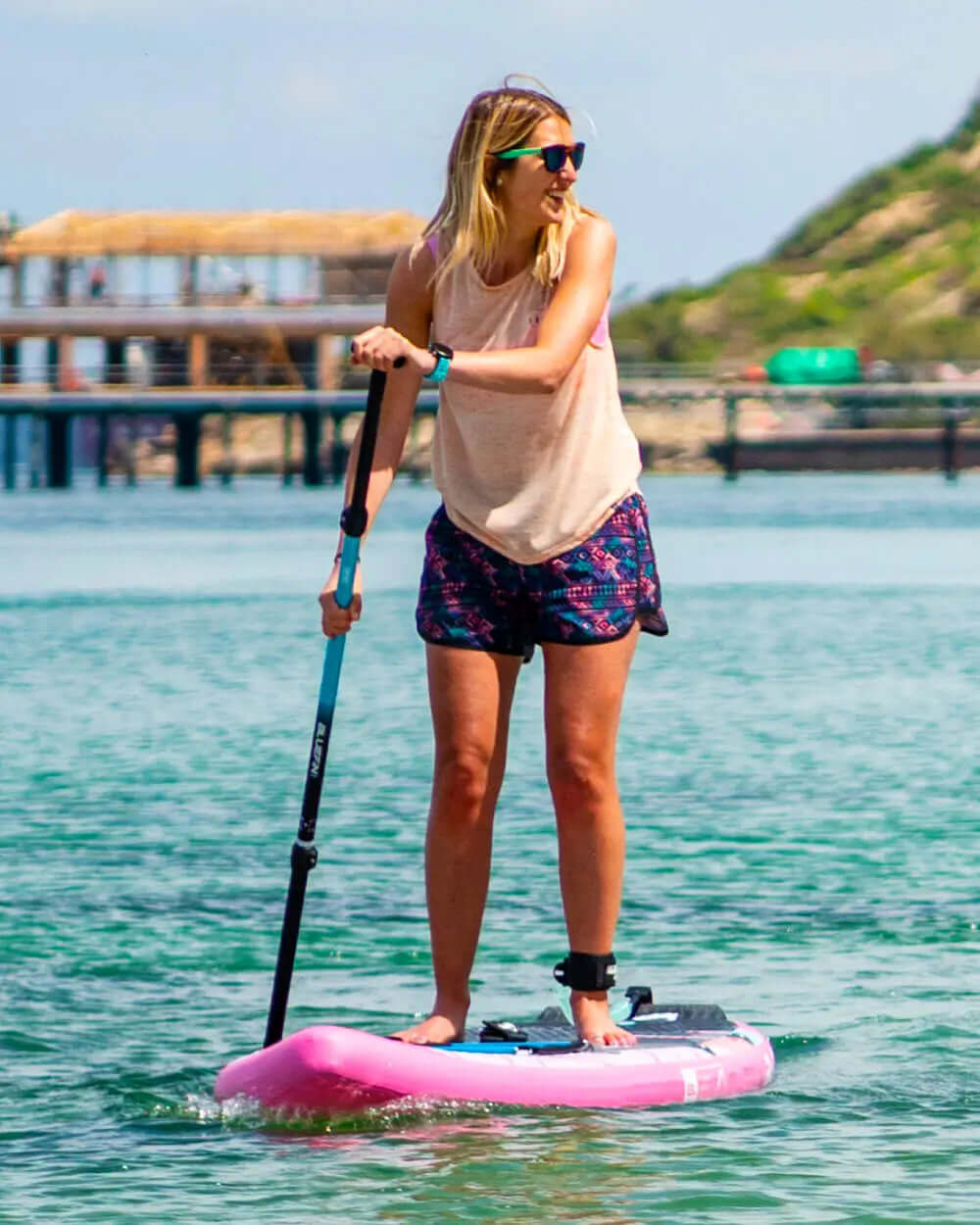 Woman paddleboarding on a pink inflatable board in clear water, wearing sunglasses and casual attire.