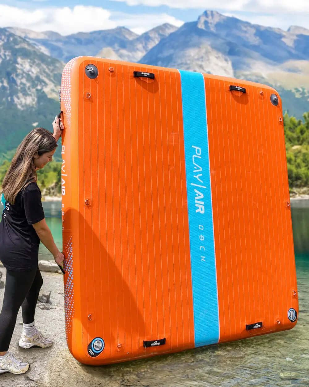 Orange inflatable dock with blue stripe, person lifting it near water and mountains in background