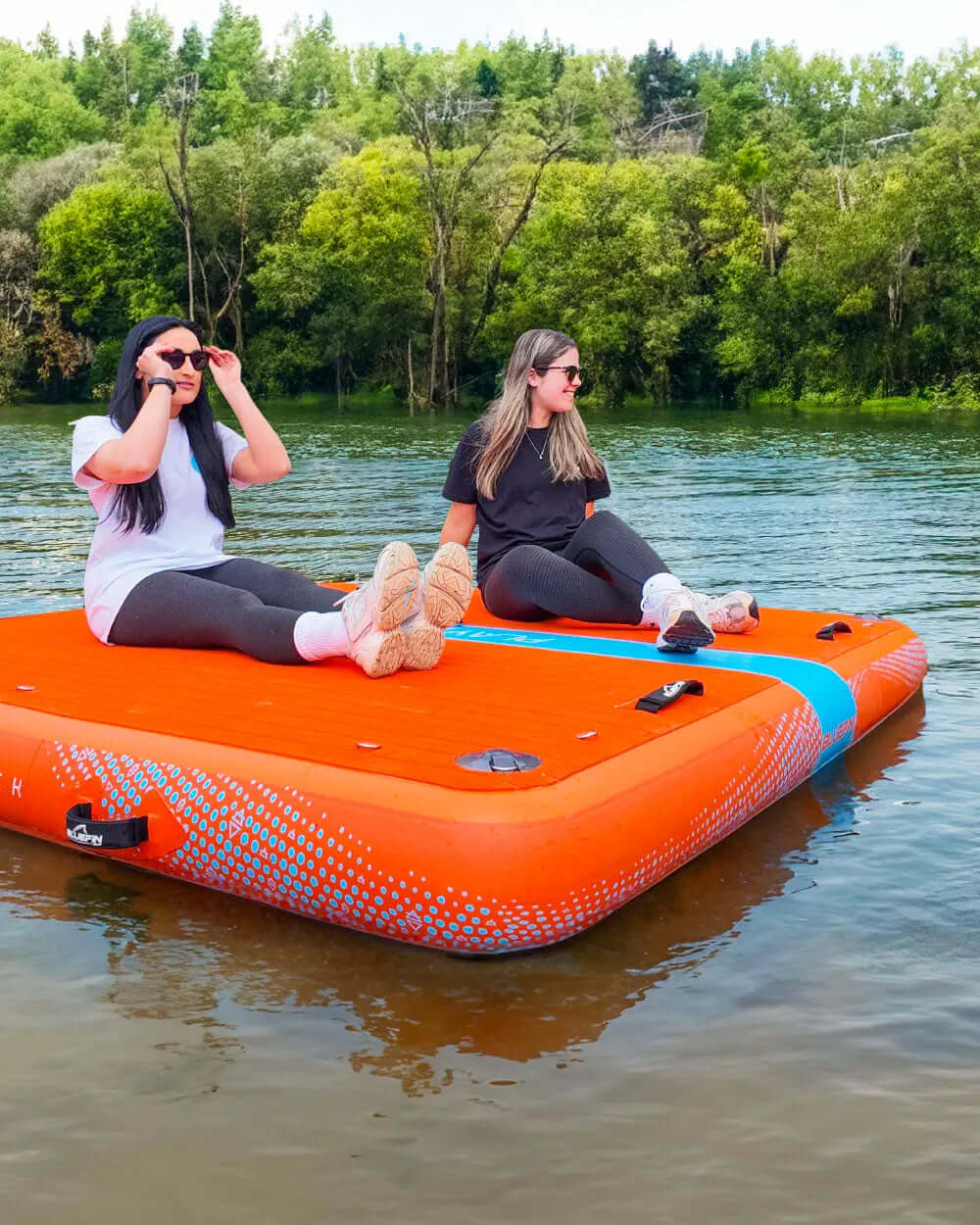 Two women sitting on an orange inflatable platform on a lake, wearing sunglasses and casual clothing.