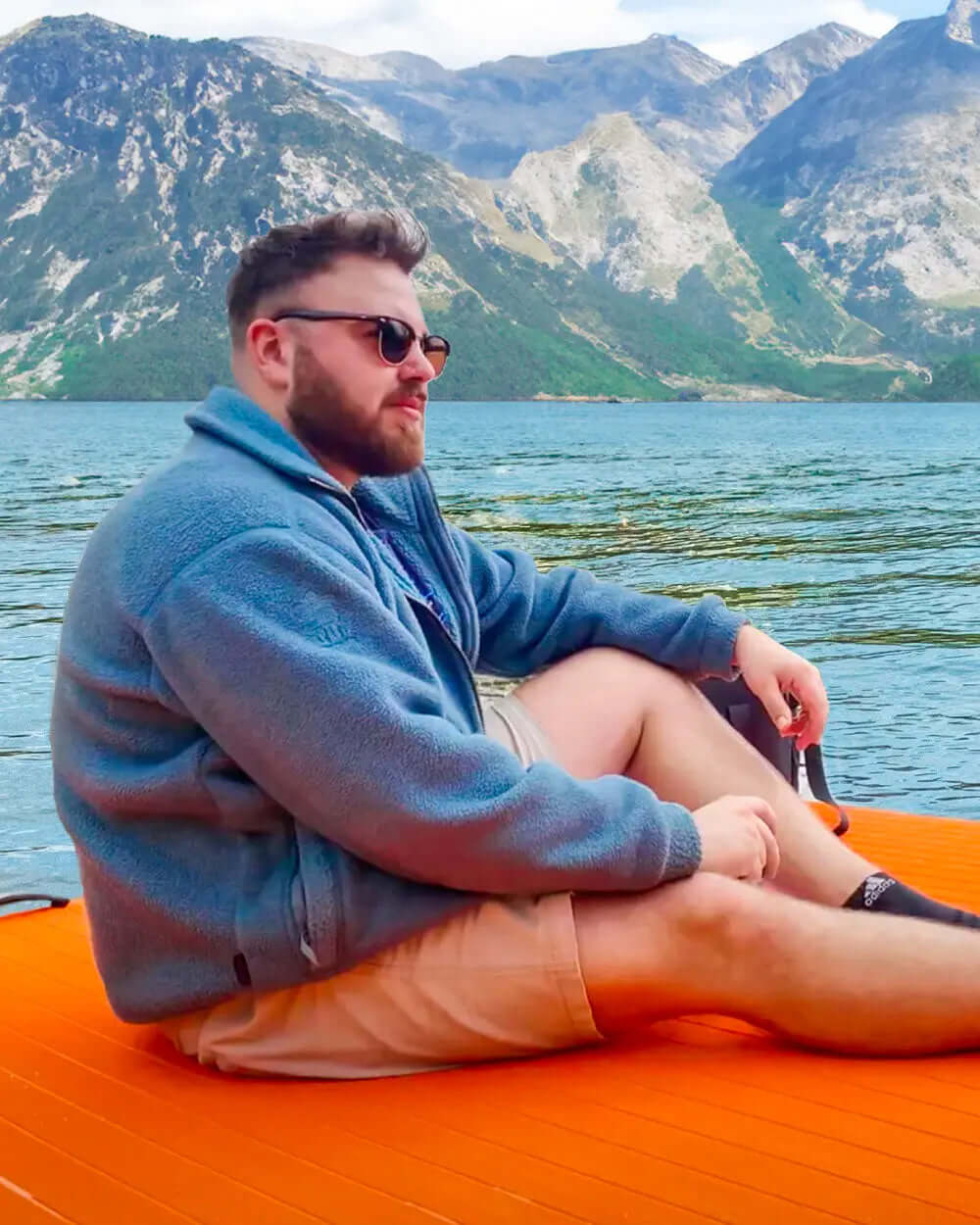Man sitting on an orange paddleboard, wearing sunglasses and a blue fleece, with mountains in the background.