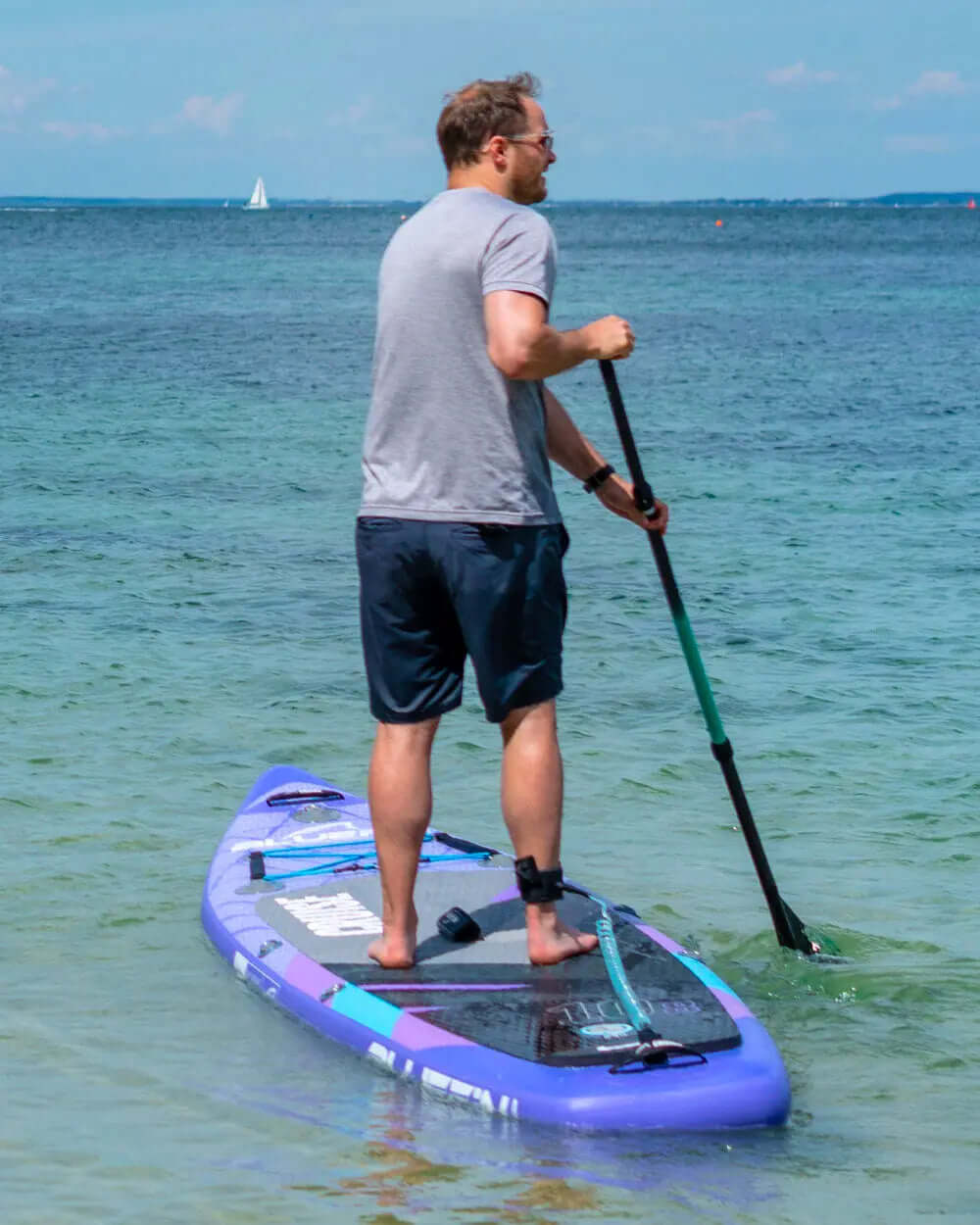 Man paddling on a purple inflatable paddleboard in clear water, wearing gray shirt and black shorts.