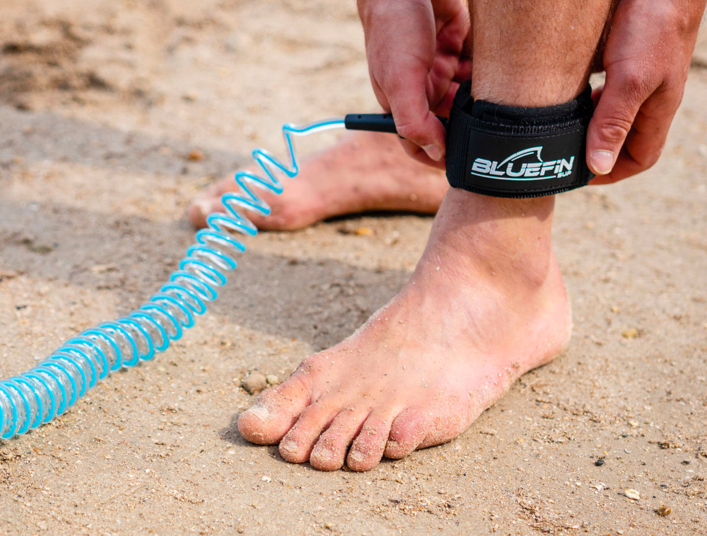 Person attaching a blue coiled leash to a black ankle strap on sandy beach